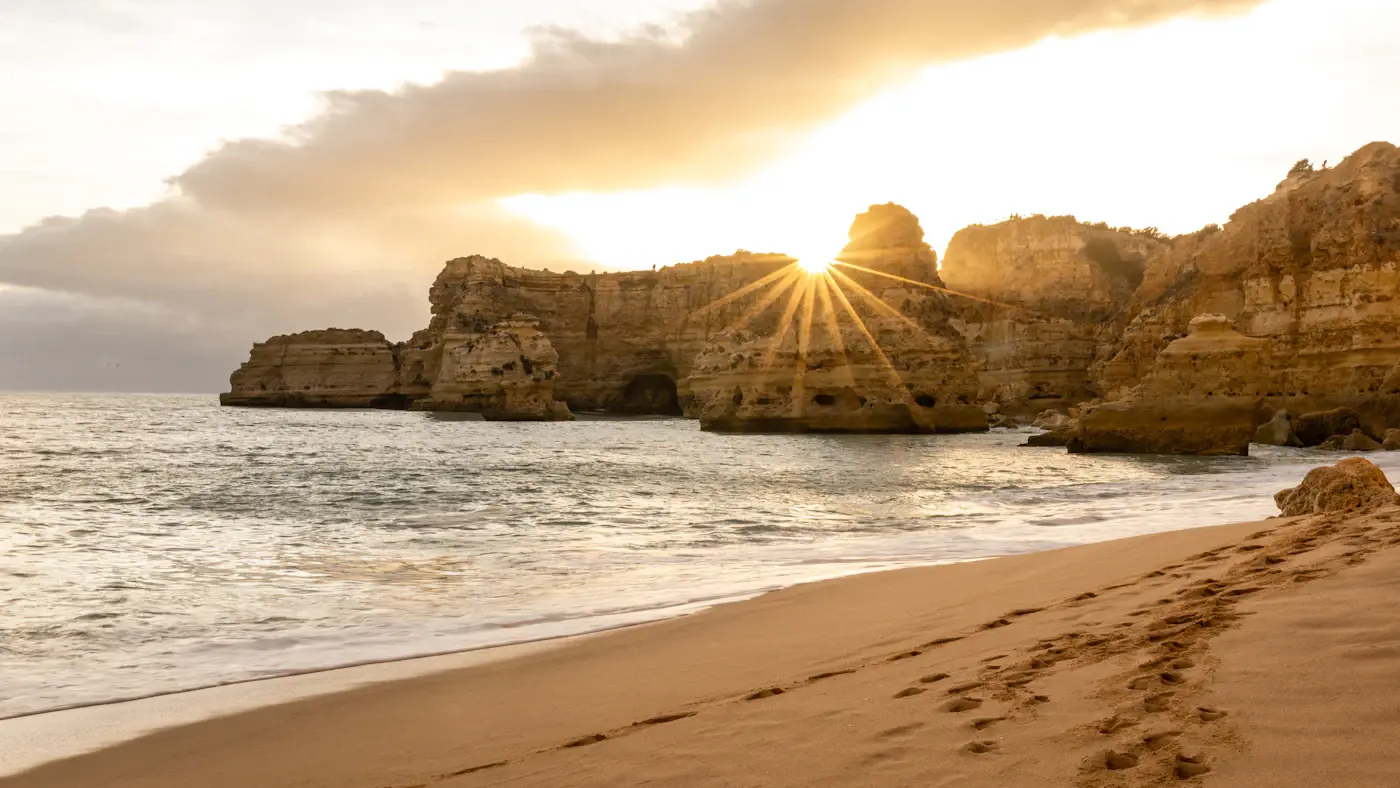 A sandy Algarve beach at sunset, the sun low behind a golden rock stack